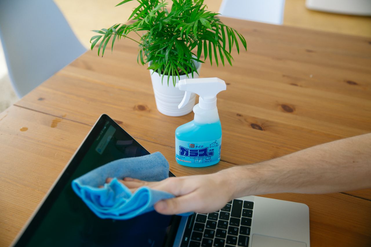 home-img Gros plan sur une main nettoyant l'écran d'un ordinateur portable avec un chiffon bleu et un vaporisateur posé sur un bureau en bois.